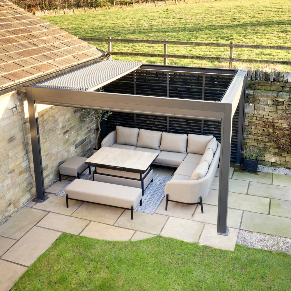 The Abierto Motorised 3m x 4m Pergola with Retractable Roof and LED Lighting in Grey creates an inviting outdoor space beside a stone wall, perfect for relaxing on your patio. Installation and delivery are included.