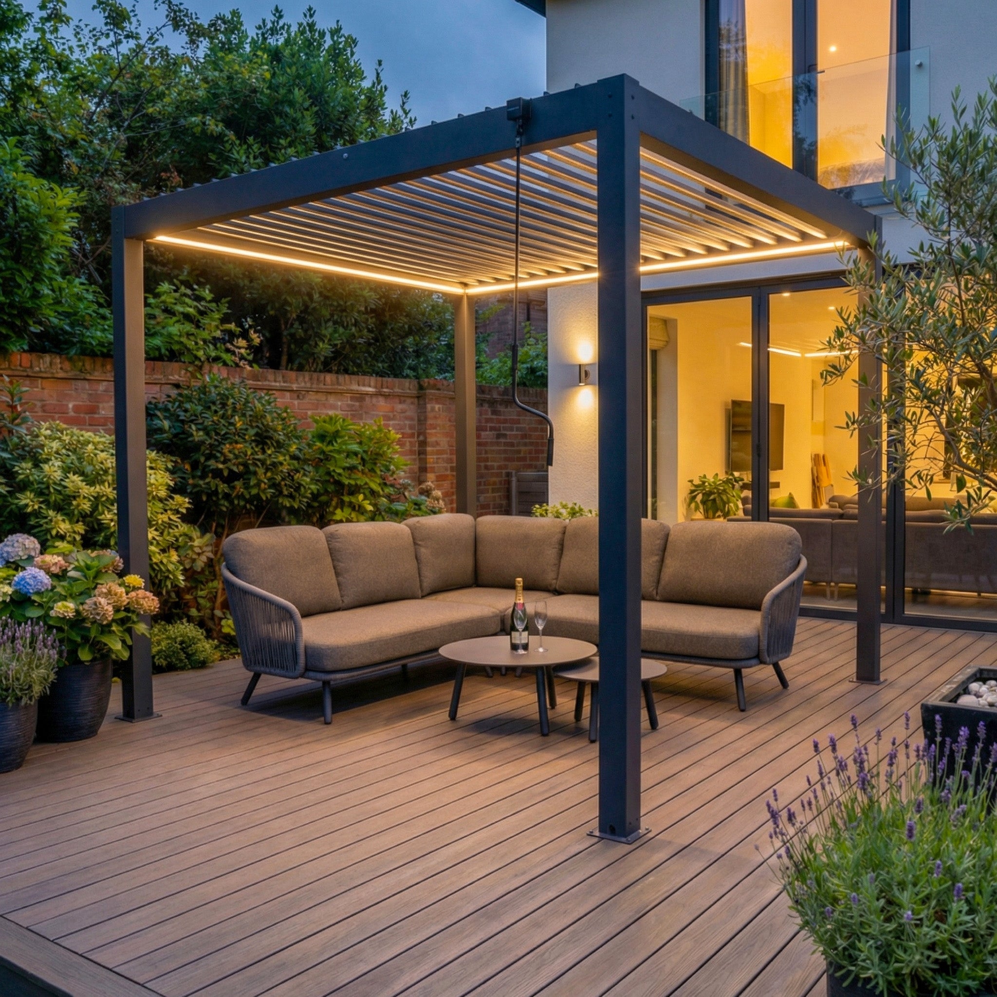 Modern patio featuring the PergoSTET Plus 3m x 3m Square Pergola (Frame Only) with integrated LED lighting in grey, paired with a cozy sectional sofa and round table, set amid a landscaped garden.