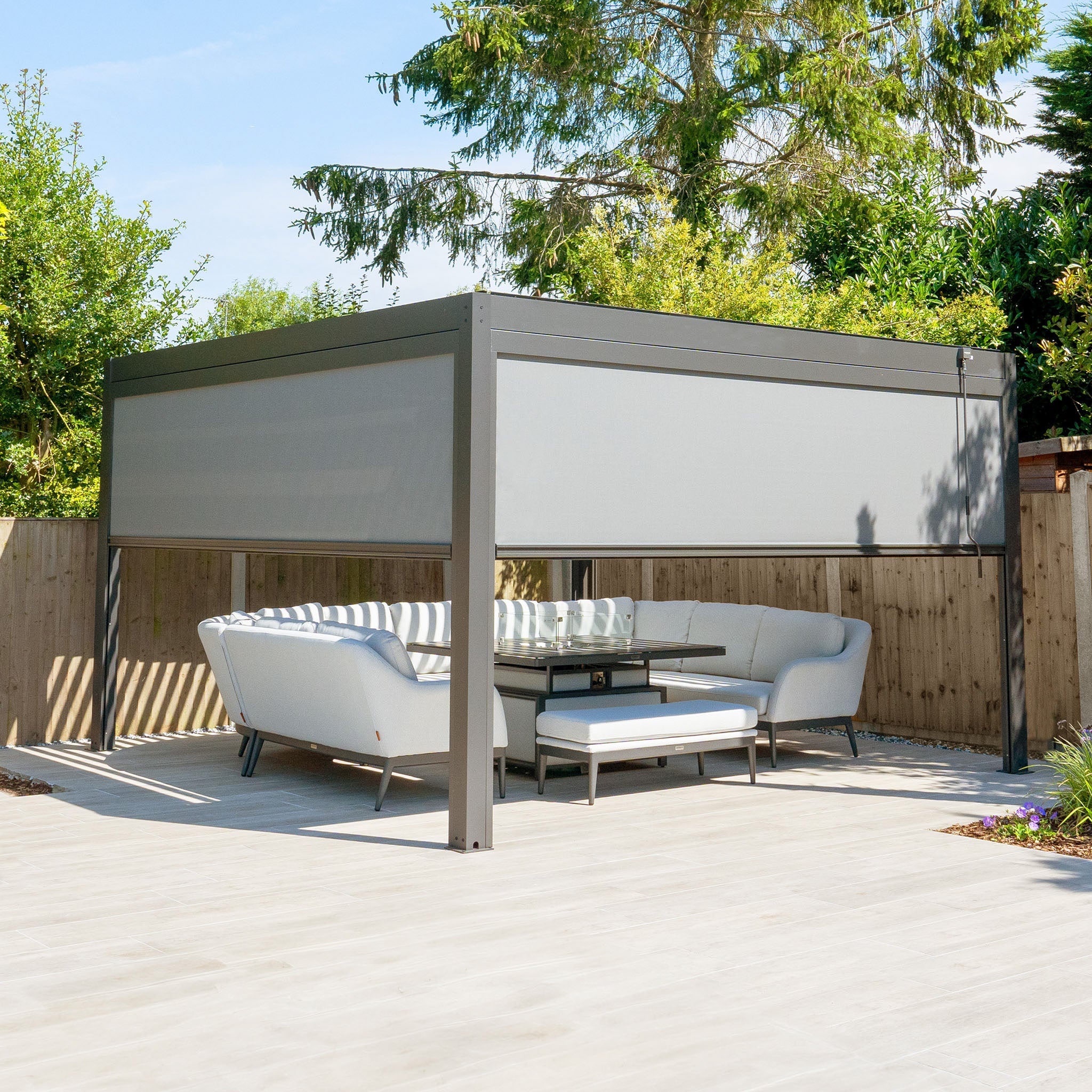 The PergoSTET Plus 4m x 4m Square Pergola features a seating area, grey design, and wooden fence backdrop for ideal outdoor living. Its subtle LED lighting enhances ambiance for evening gatherings.