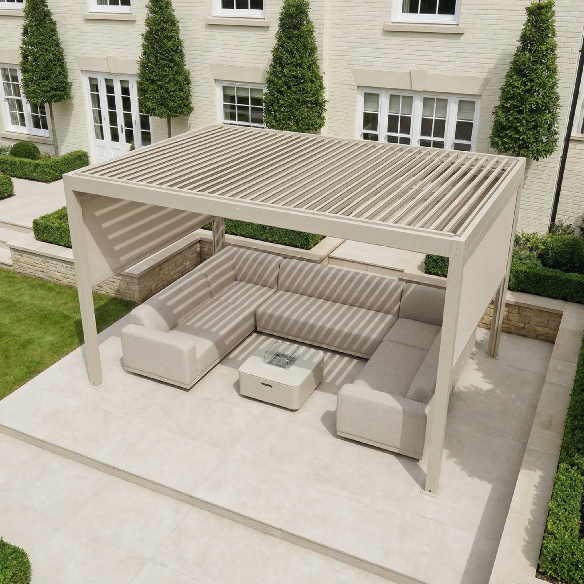 PergoSTET Plus 3m x 4m Rectangular Pergola with 3 manual side blinds and LED lighting in Latte creates a stylish modern patio setup, complete with sectional sofa and table, outside a white house with trimmed trees.