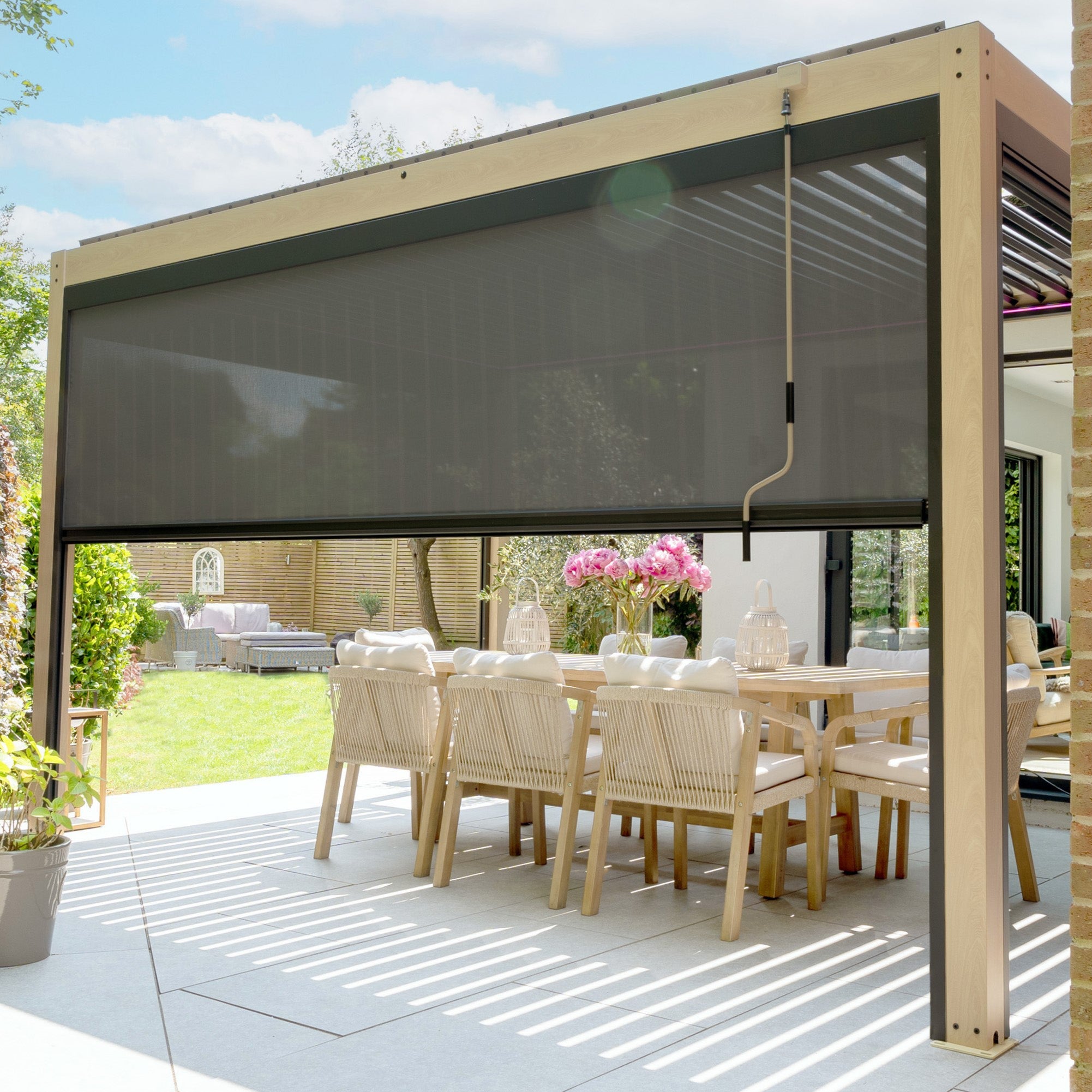 The PergoSTET Plus 3m x 4m Rectangular Pergola, featuring adjustable louvres, LED lighting, and a wood effect finish, provides versatile coverage for an outdoor dining area with wooden chairs and a table.